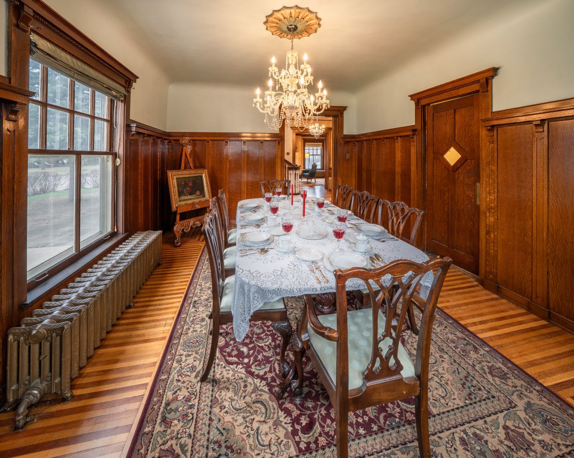 A dining room with a long table set for six, surrounded by wooden chairs, a chandelier, and a large window with a view.