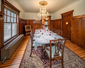 A dining room with a long table set for six, surrounded by wooden chairs, a chandelier, and a large window with a view. - Perry Mansion | Dutch Colonial (Sioux Falls)