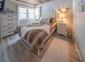 Ein Schlafzimmer mit einem großen Bett, einem Nachttisch mit Lampe, einer Kommode und einem Fenster mit Blick.