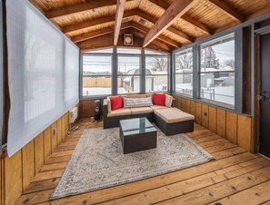 Ein gemütliches Zimmer mit Holzbalkendecke, großen Fenstern, einer Eckcouch und einem Glastisch.