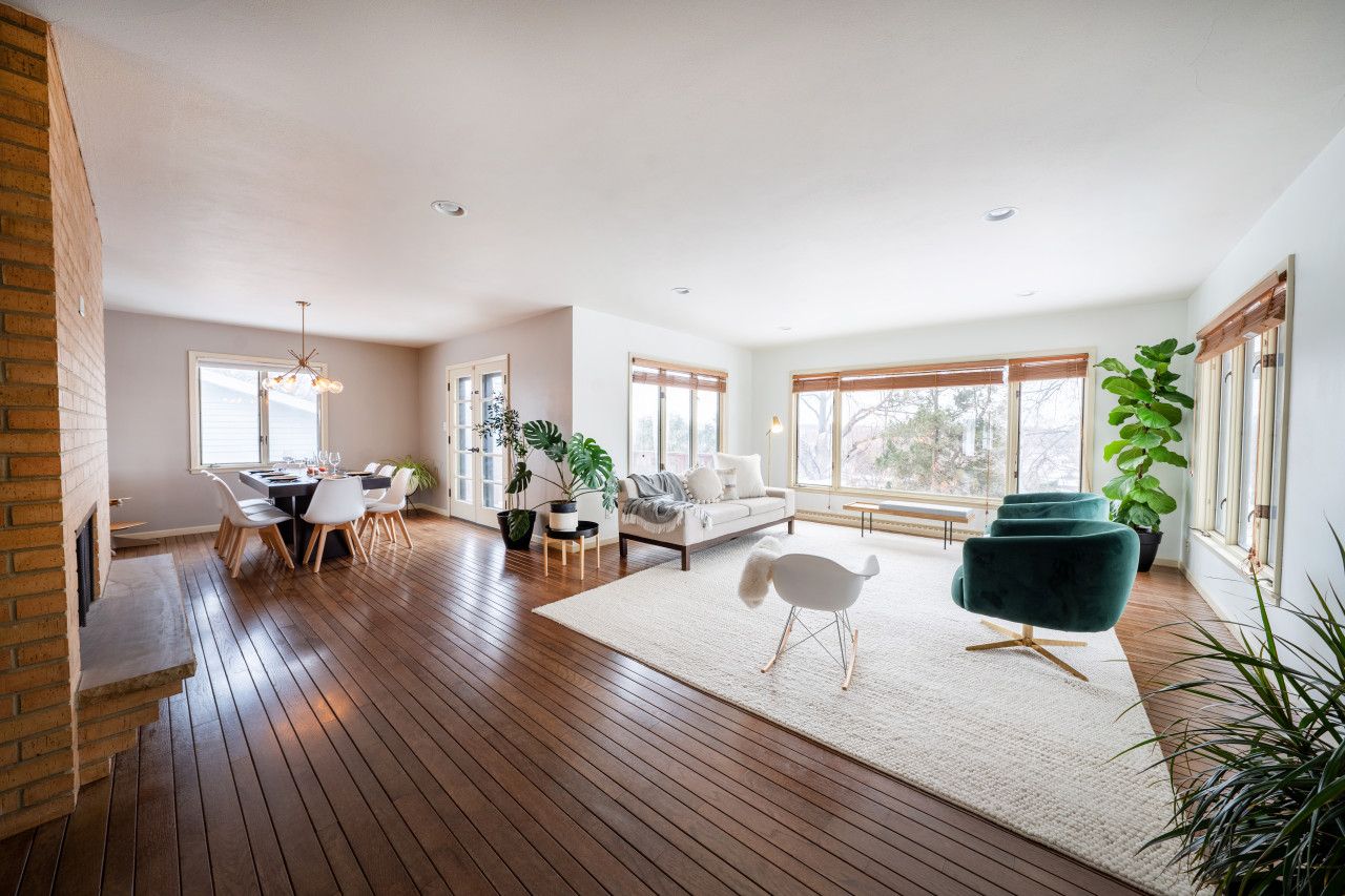 A spacious living area with a wooden floor, a dining table with chairs, a sofa, and a large window with blinds.