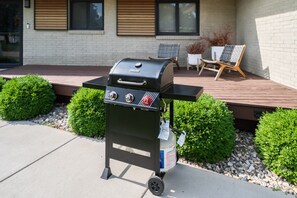 A portable gas grill with a side table and a gas tank, placed on a concrete surface with a wooden deck and potted plants in the background.
