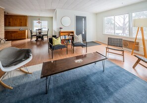 A spacious living area with a dining table, chairs, a bookshelf, and a large window providing natural light.