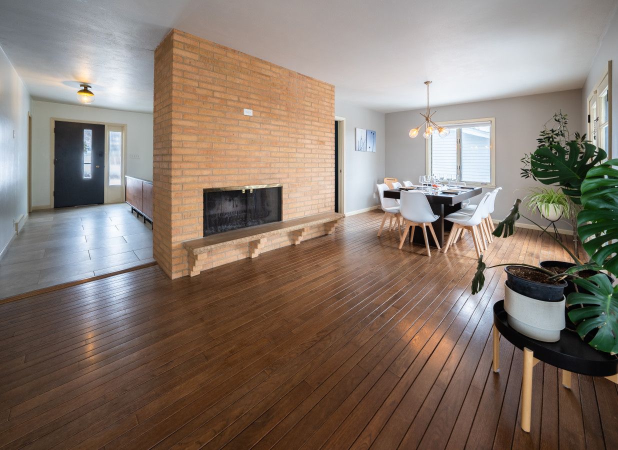 A spacious living area with a brick fireplace, wooden flooring, a dining table with chairs, and potted plants.