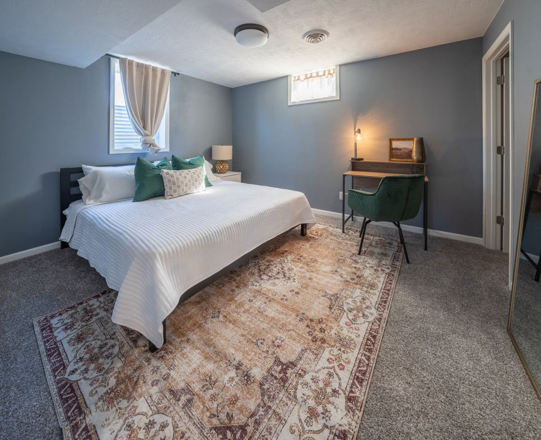Un dormitorio con cama, escritorio, silla, alfombra y ventana con cortinas.