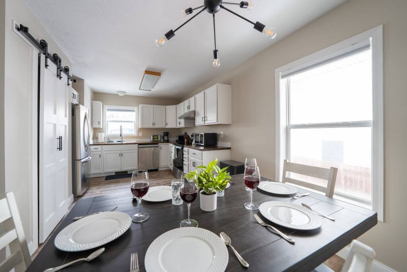 A modern kitchen with white cabinets, a dining table set for two with plates, glasses, and cutlery, and a potted plant on the table.
