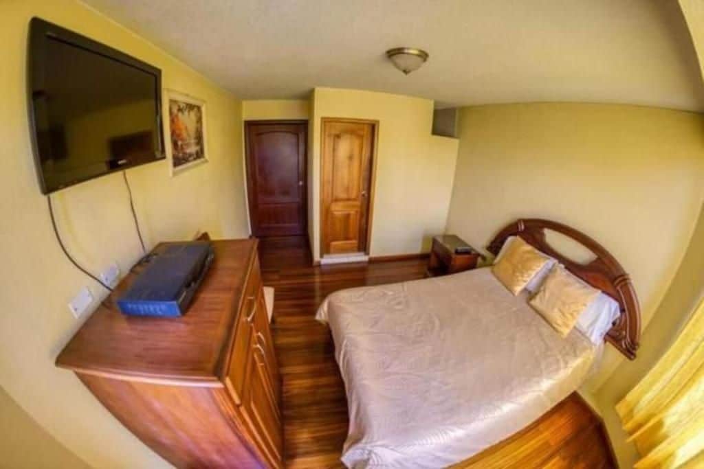 A bedroom with a bed, a wooden dresser, a television mounted on the wall, and a door leading to another room.