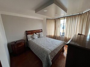 A bedroom with a bed, a nightstand, a dresser, and a window with curtains. - Hotel Rocio (Riobamba)