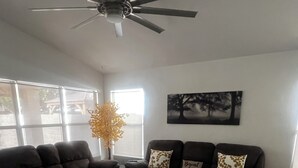Una sala de estar con dos sofás oscuros, un tapete estampado, un ventilador de techo y una ventana con vistas a árboles.