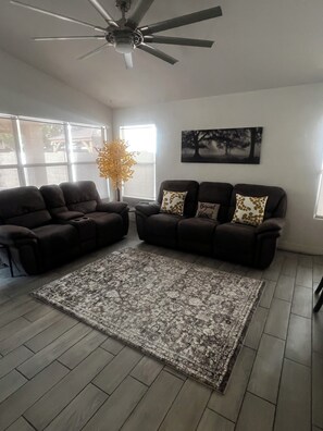 A living room with two dark sofas, a patterned rug, a ceiling fan, and a window with a view of trees. - Our home comfortably sleeps up to 6 guests and features 2 bedrooms+bonus room. (Avondale)