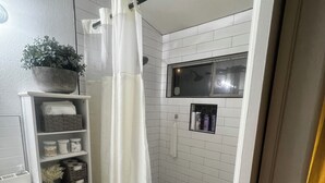 Un baño con ducha, un lavamanos blanco, un estante con artículos de aseo y una planta en maceta.