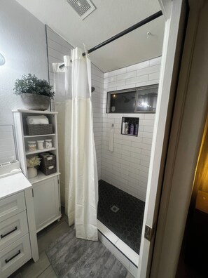 A bathroom with a shower, a white sink, a shelf with toiletries, and a potted plant. - Our home comfortably sleeps up to 6 guests and features 2 bedrooms+bonus room. (Avondale)