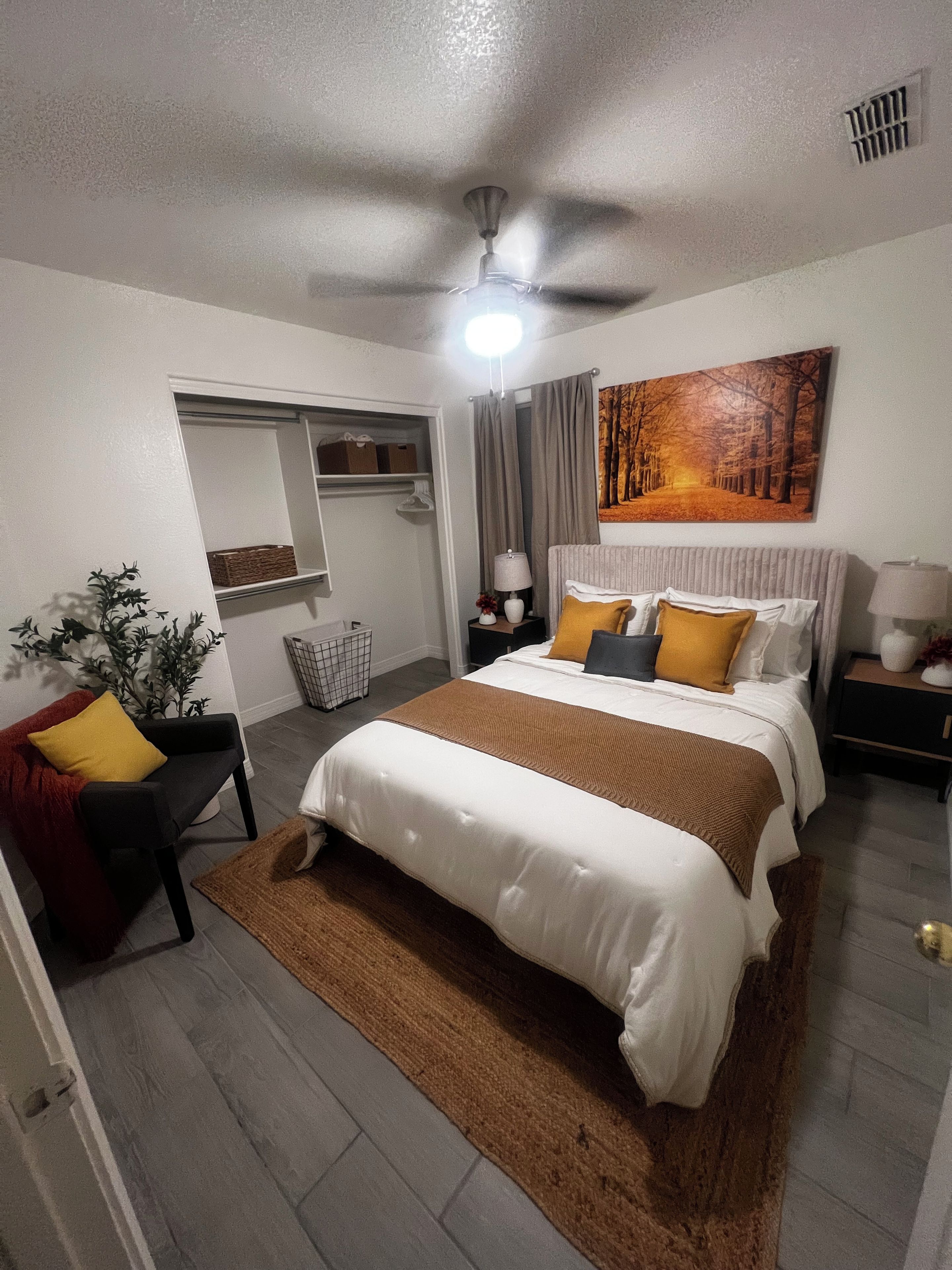 Quarto com cama, mesas de cabeceira, ventilador de teto, um quadro e porta que dá para um armário.
