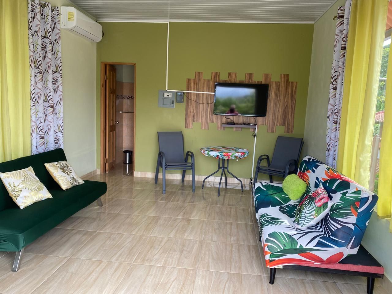 Ein Wohnzimmer mit einem grünen Sofa, einem farbenfrohen Sofa, einem kleinen Tisch und einem an der Wand befestigten Fernseher.