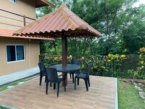 An outdoor patio with a round table and chairs, a metal umbrella, and a garden with yellow flowers. - Private Oasis Villa: Pool, BBQ Area & Lush Nature - Family Friendly (Provincia de Panamá Oeste, Corregimiento de Guayabito)