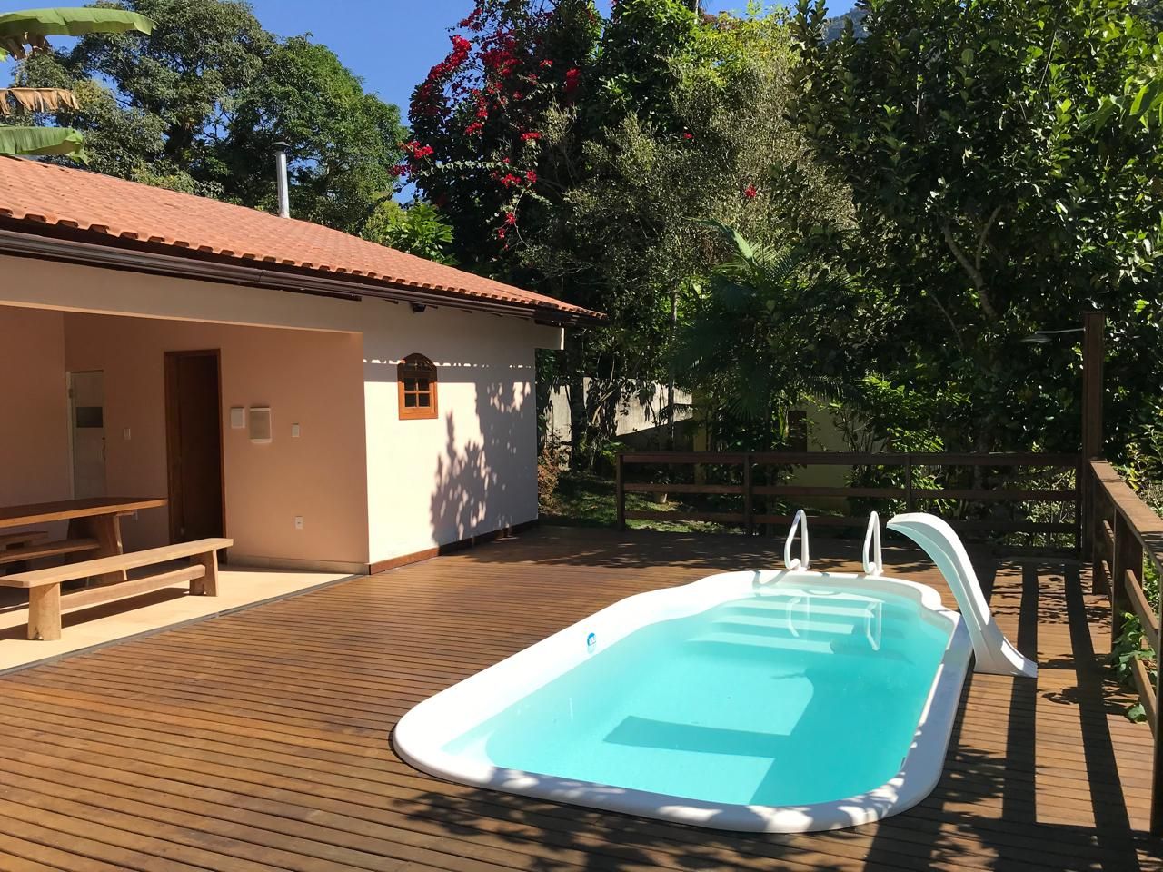 Piscina com deck de madeira, um pequeno edifício com telhado vermelho e vegetação exuberante ao fundo.