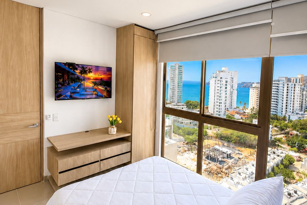 A bedroom with a large bed, a mounted TV, a wooden cabinet, and a window with a city view.