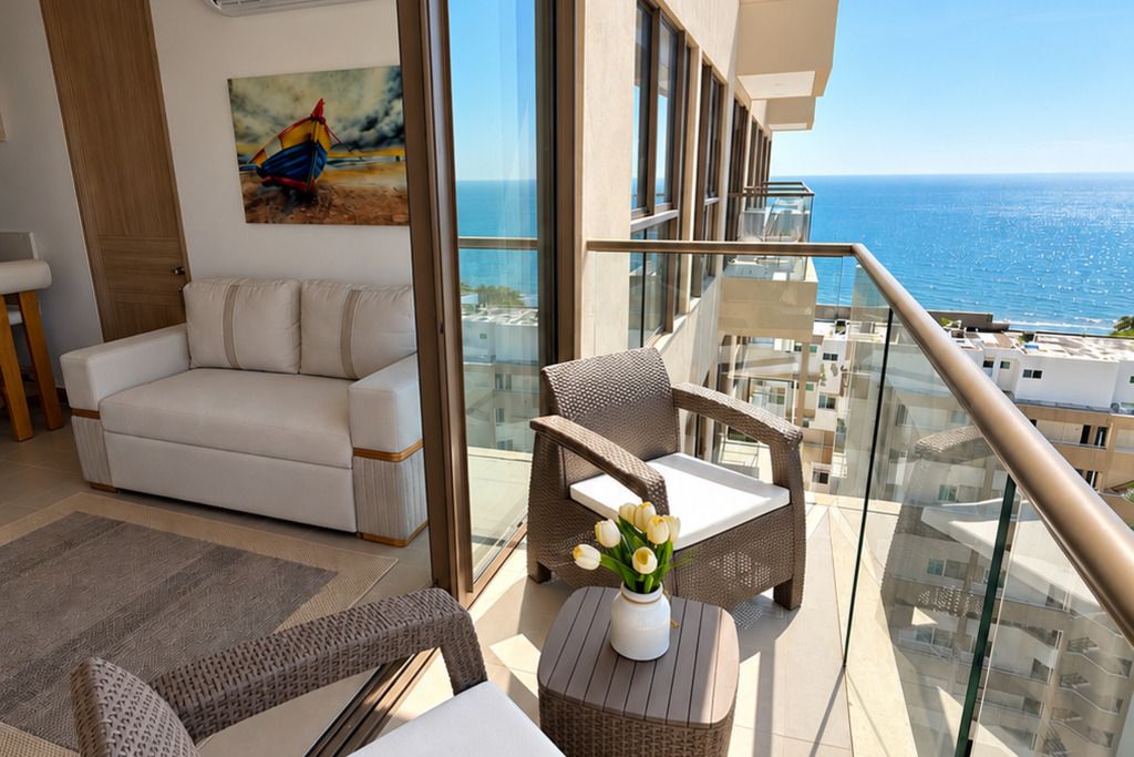 A balcony with a view of the sea, a white sofa, wicker chairs, and a small table with flowers.