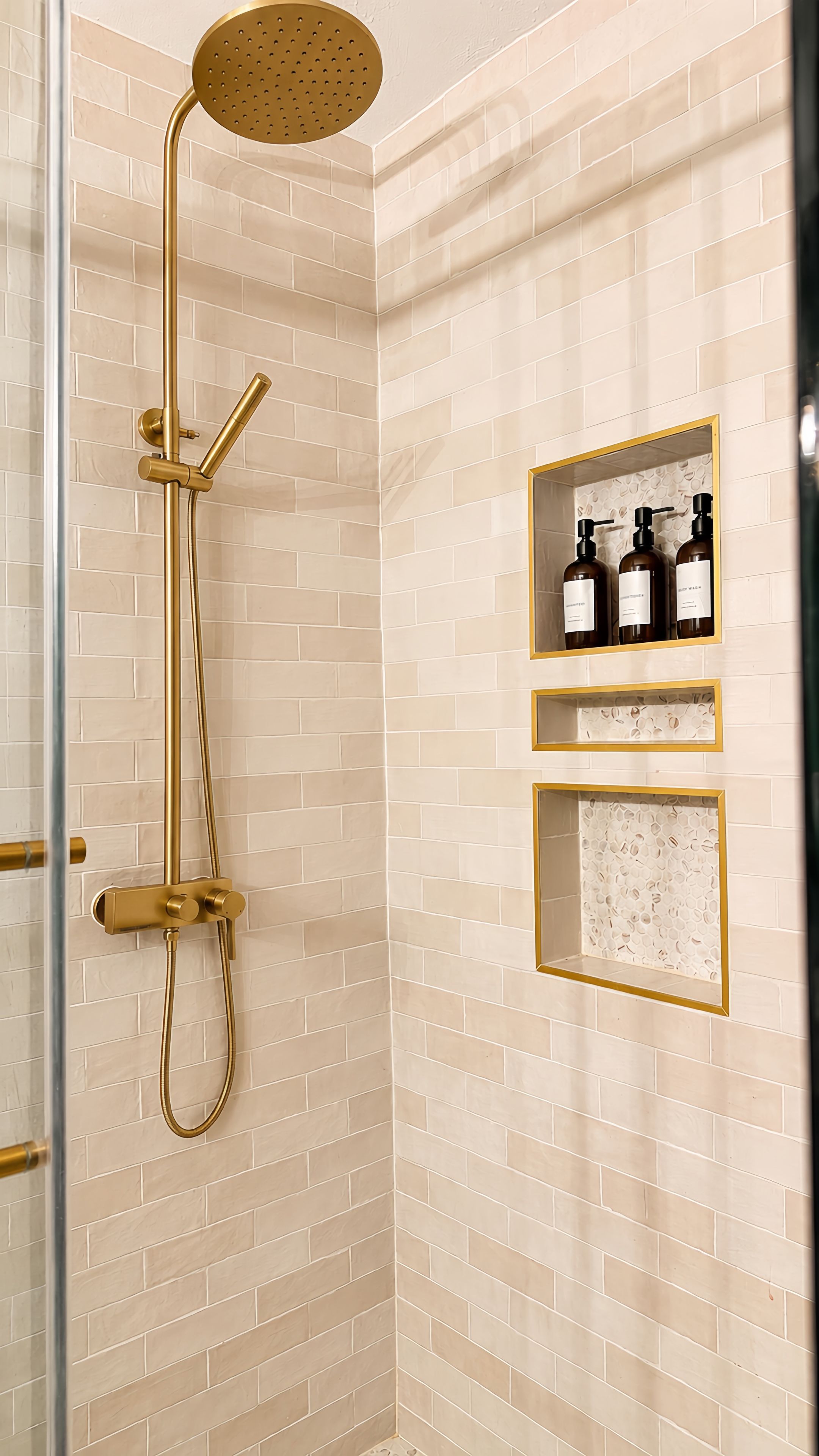 Ducha con cabezal y manguera dorados, puerta de cristal, pared con azulejos color beige y dos repisas con botellas.