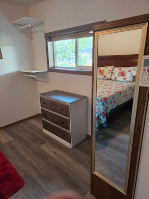 Un dormitorio con un aparador de madera, una cama con ropa de cama floral, una ventana con vistas y un espejo con reloj digital.