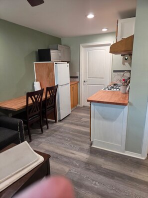 Cocina compacta con refrigerador, microondas y estufa, zona de comedor con mesa y sillas, y un pequeño rincón de descanso con sofá y sillón.
