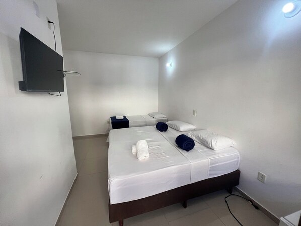 A single bed with white bedding and two pillows, a bedside table with a lamp, and a flat-screen TV mounted on the wall.
