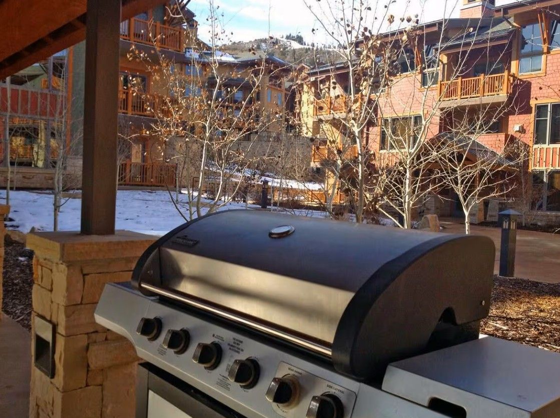 An outdoor grill with a stone structure, a wooden bench, and a view of a building with balconies and a snow-covered ground.