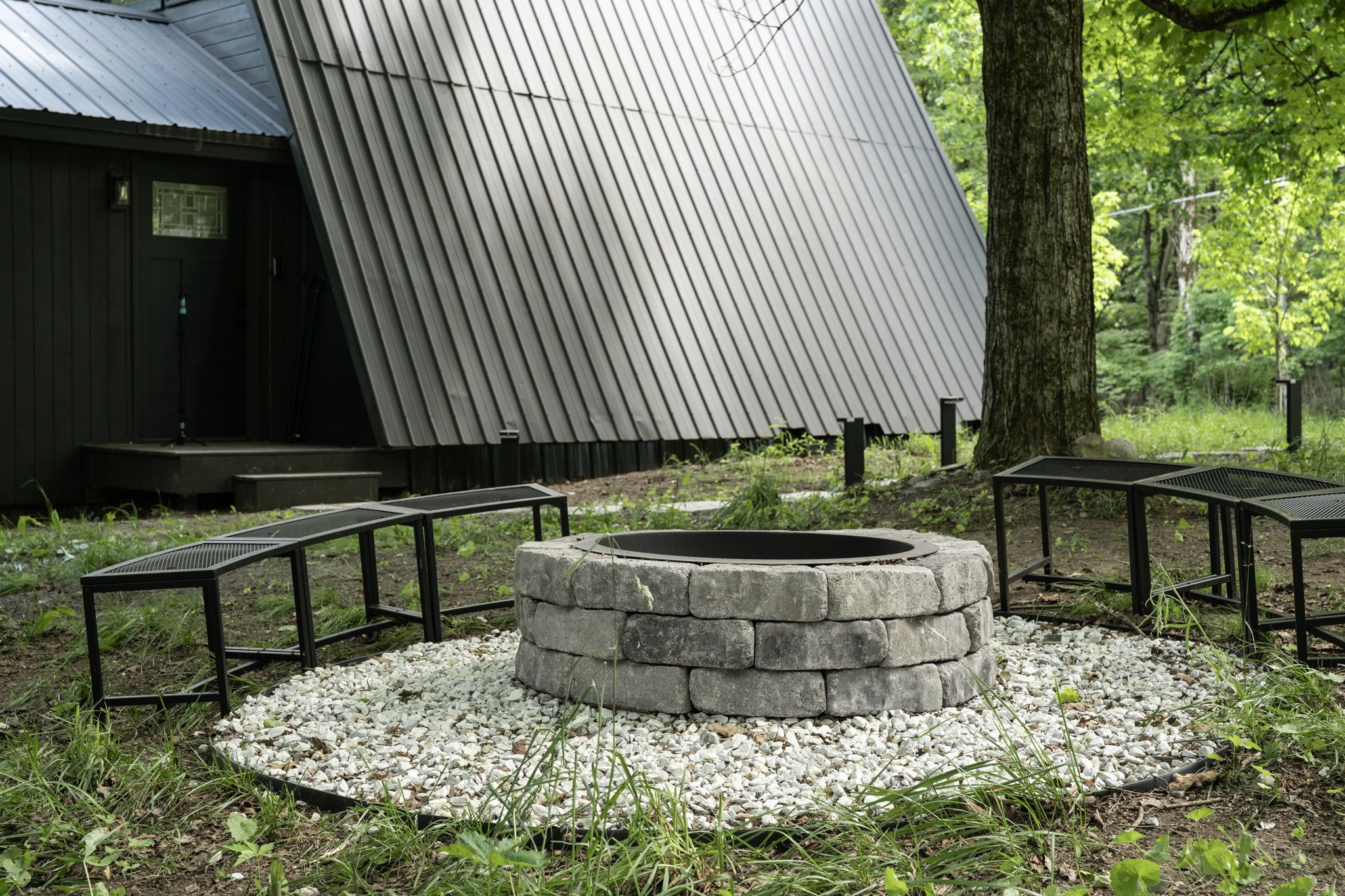 A stone fire pit surrounded by gravel, with metal benches and a wooden structure in the background.