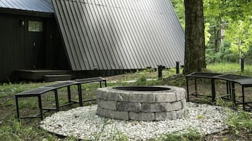 A stone fire pit surrounded by gravel, with metal benches and a wooden structure in the background.