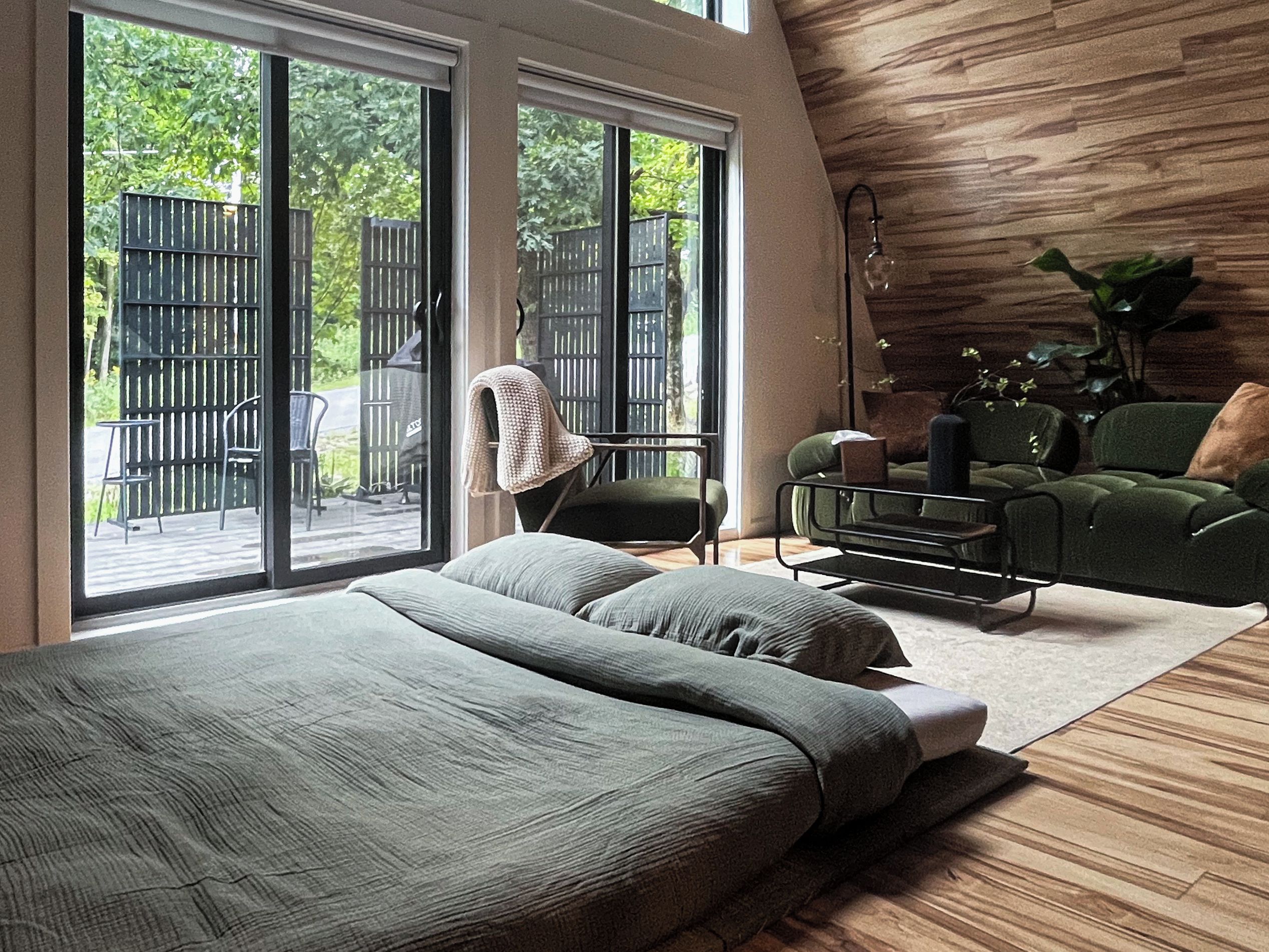 A modern bedroom with a large bed, a green sofa, and a chair. There is a sliding glass door leading to an outdoor area with a wooden deck and chairs.