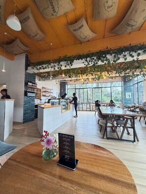 Interior de una cafetería con techo de madera, plantas colgantes, una barra y un atril para el menú.