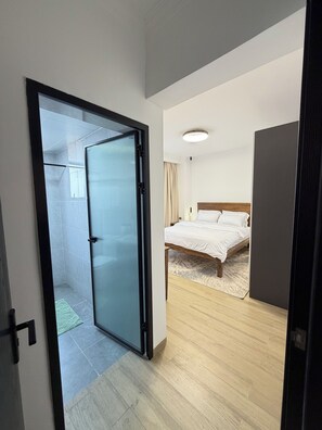 Un dormitorio moderno con cama, suelo de madera y puerta de cristal que da a la zona de ducha.