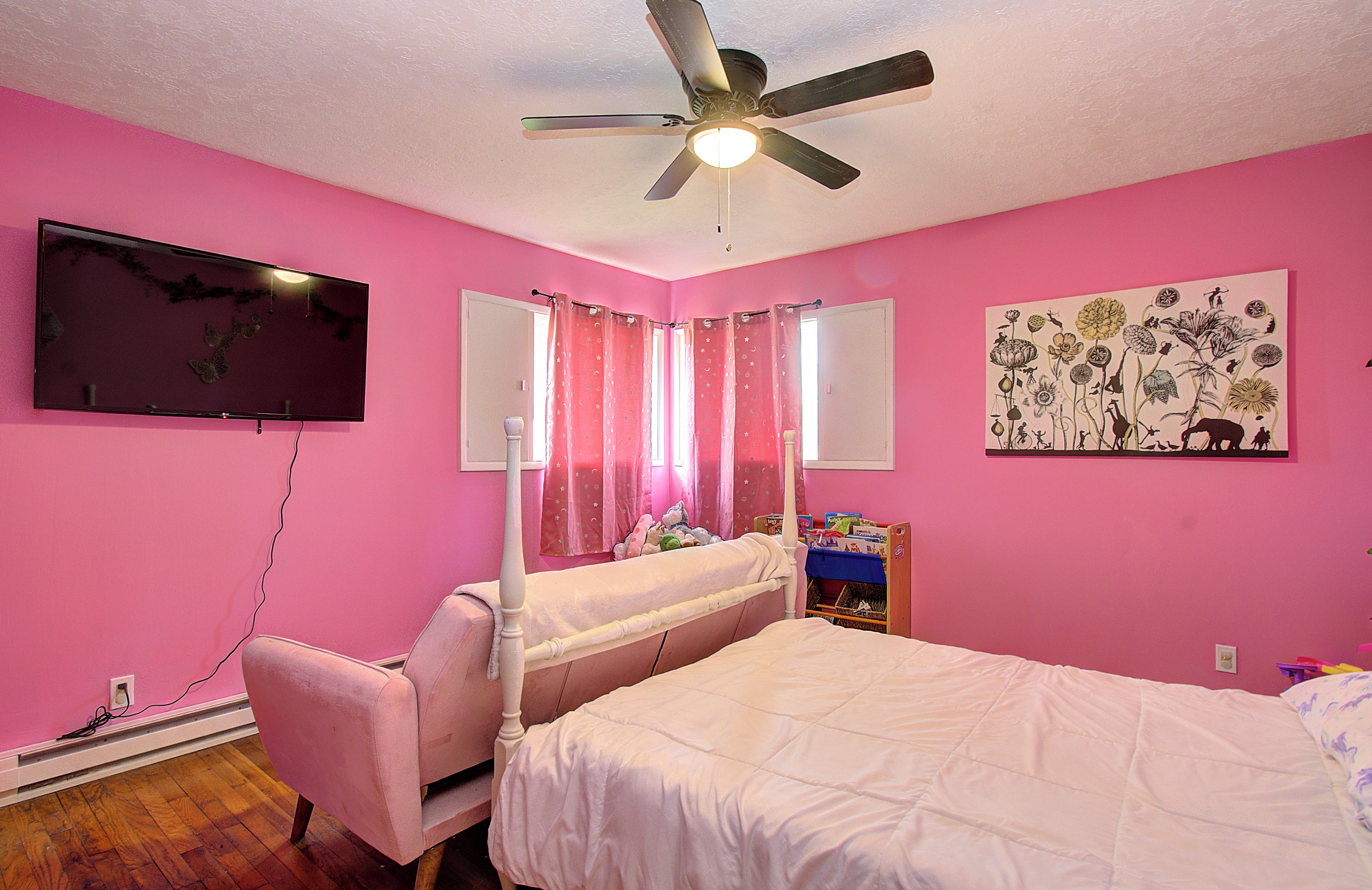 Un dormitorio decorado en tonos rosados, con cama, silla, ventilador de techo, televisor y un cuadro floral en la pared.