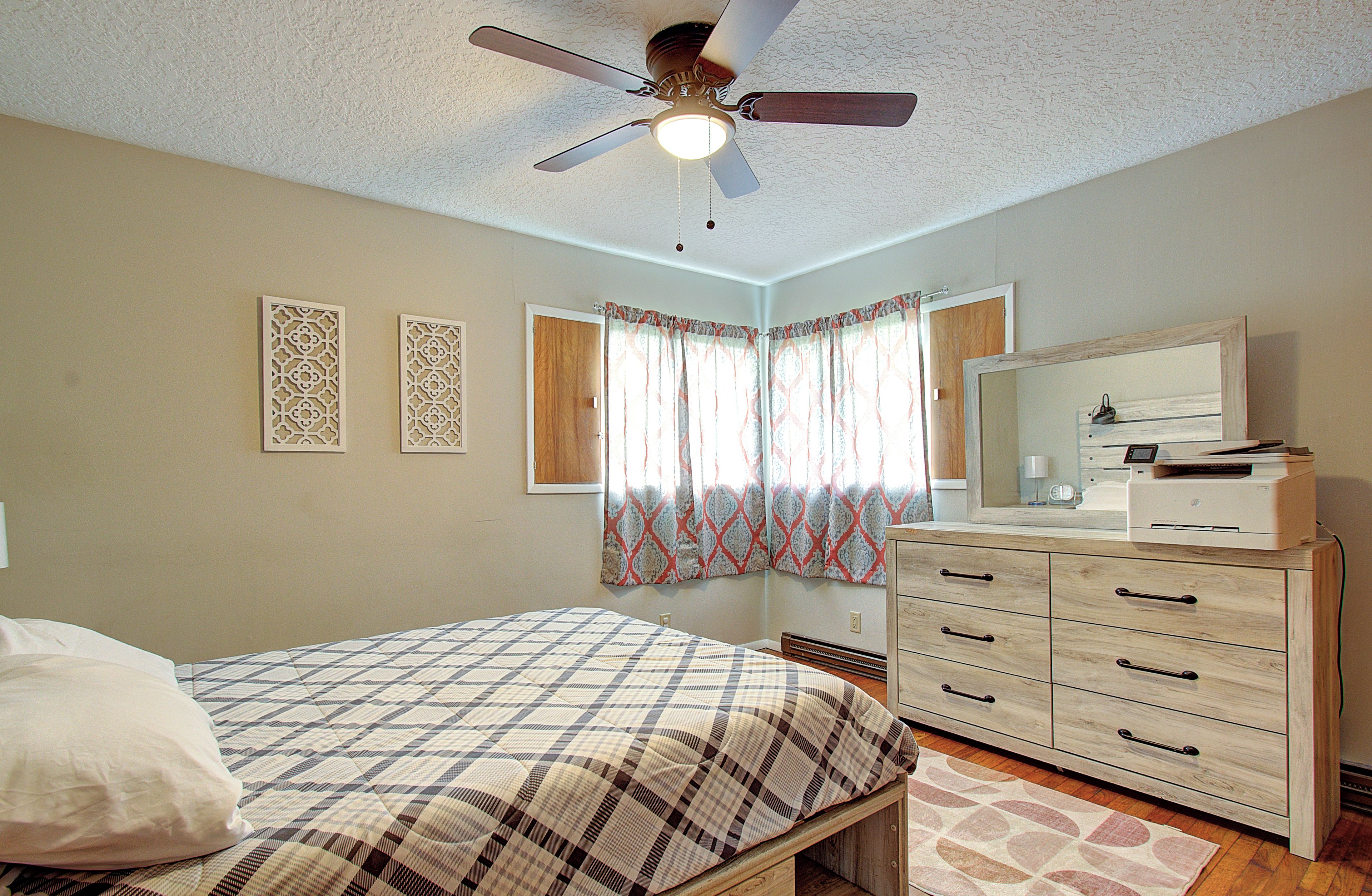 Quarto com cama, penteadeira, ventilador de teto e janela com cortinas.