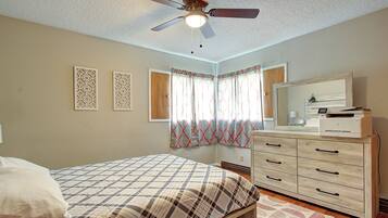Quarto com cama, penteadeira, ventilador de teto e janela com cortinas.