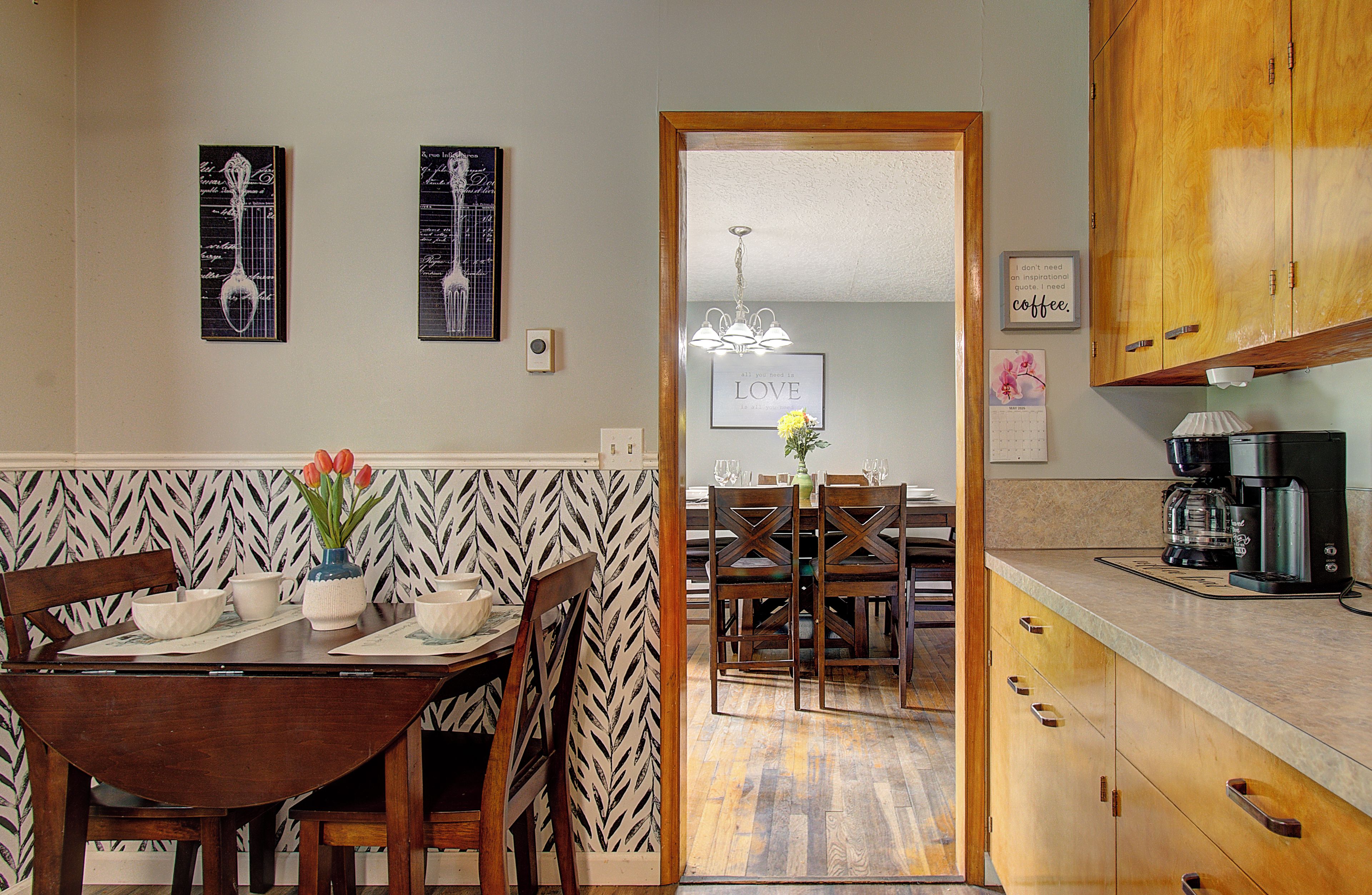 A dining area with a wooden table and chairs, a vase with flowers, and a kitchen with cabinets and a coffee maker.