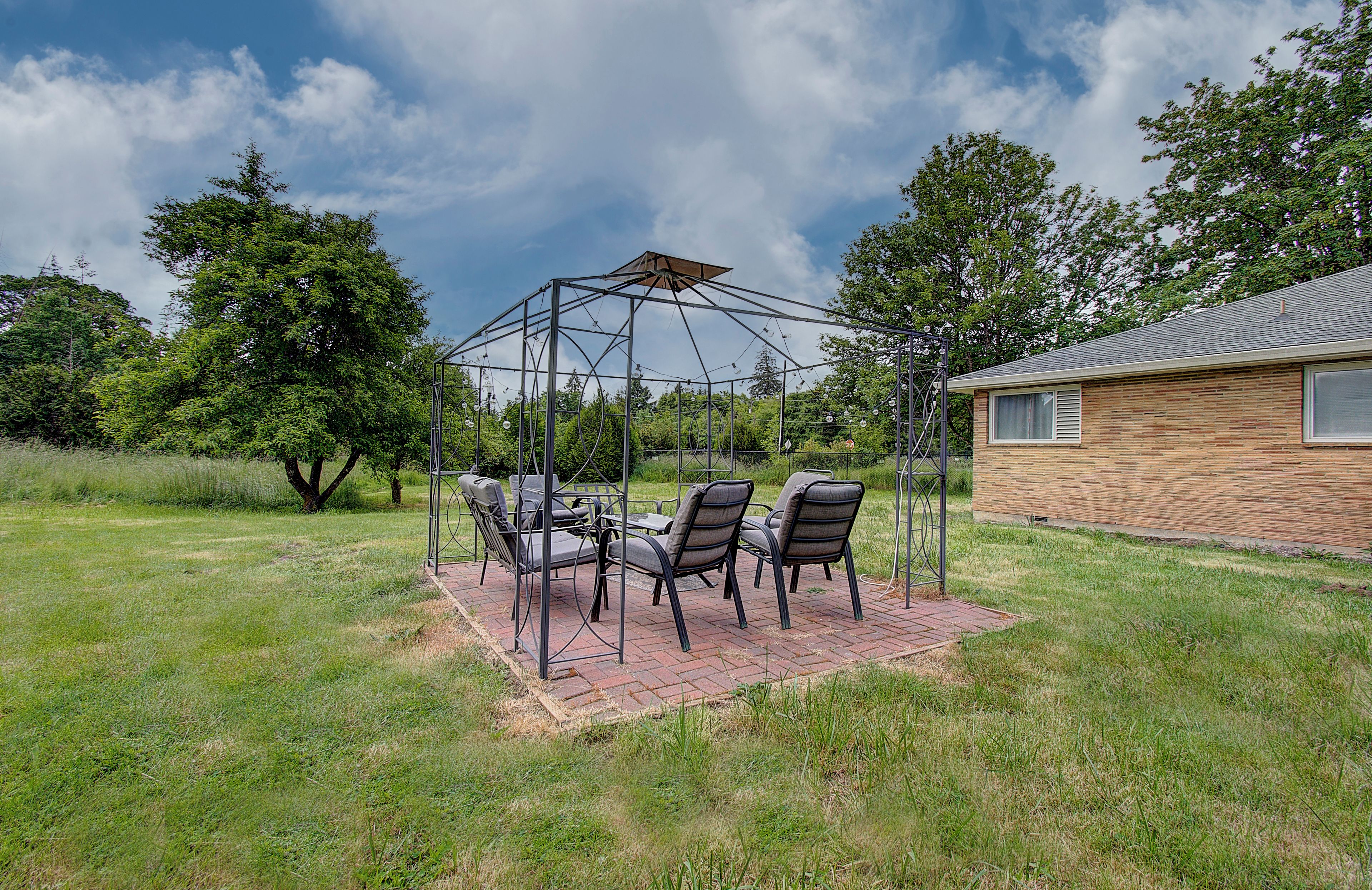 Uma área de estar coberta ao ar livre, com cadeiras e uma mesa, rodeada por um campo gramado e árvores, com uma casa ao fundo.
