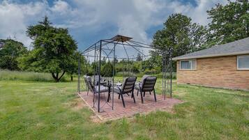 Uma área de estar coberta ao ar livre, com cadeiras e uma mesa, rodeada por um campo gramado e árvores, com uma casa ao fundo.