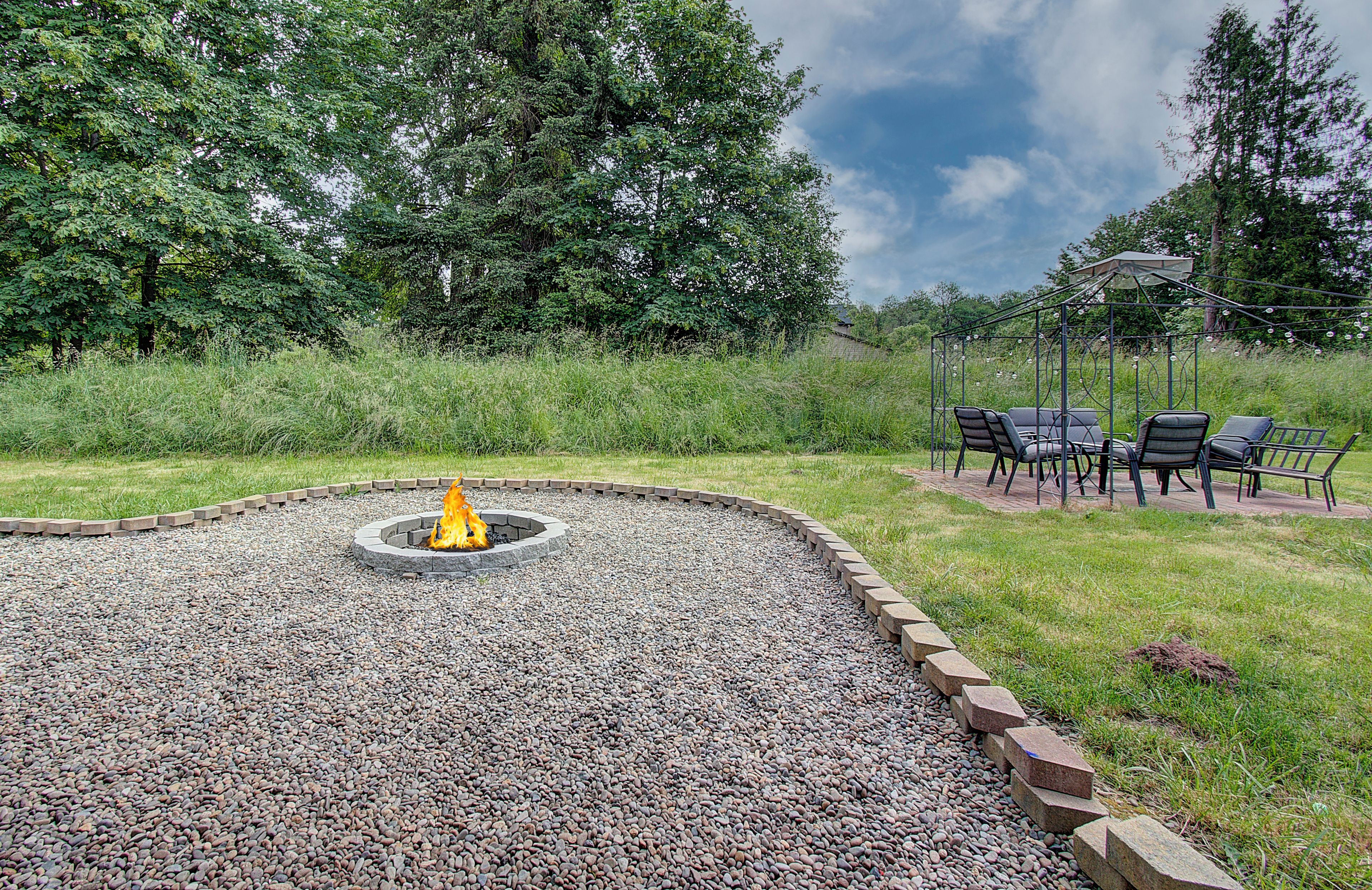 A circular fire pit surrounded by gravel, with a grassy area and a gazebo with chairs in the background.