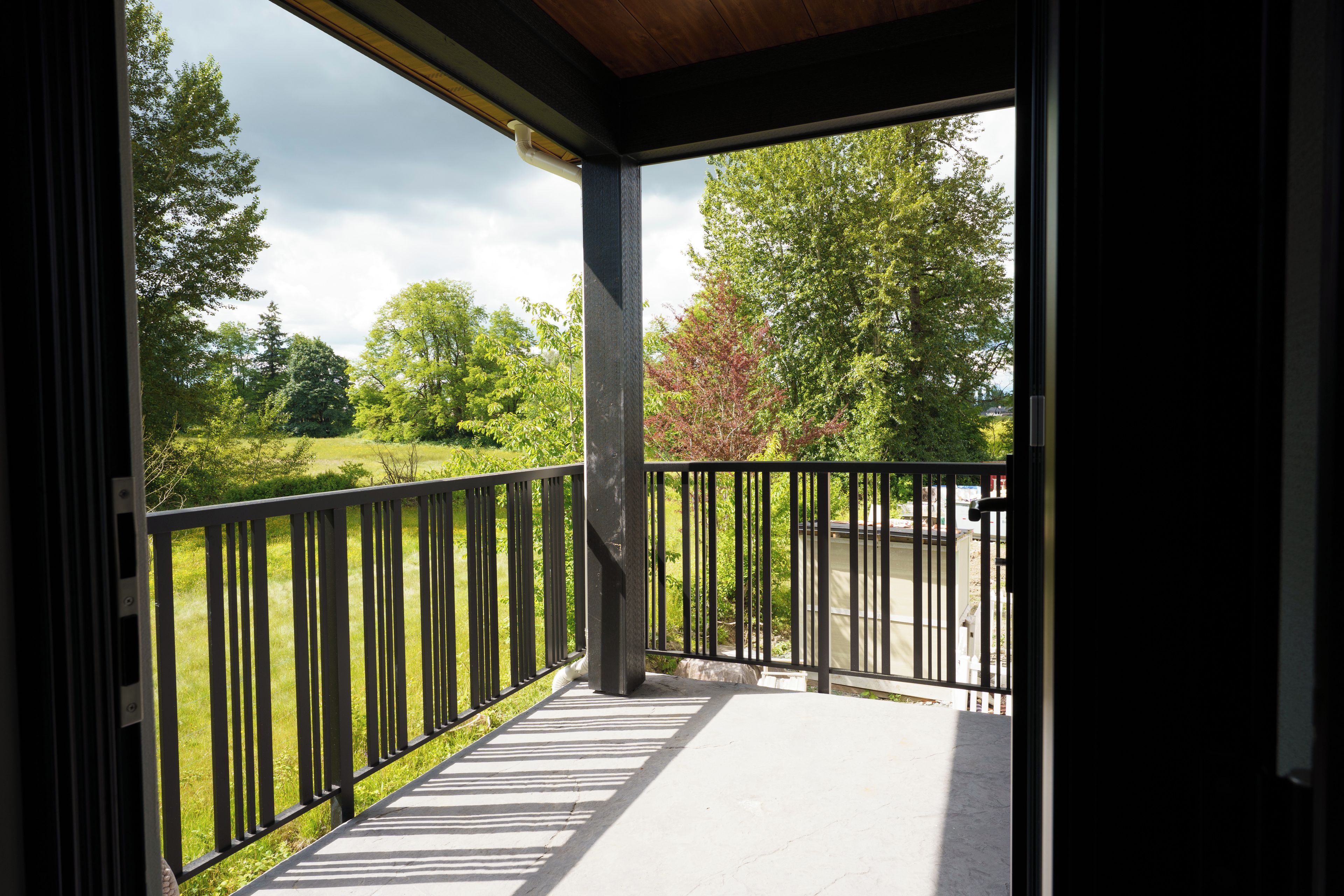 Een overdekt terras met een zwarte metalen balustrade, een betonnen vloer en uitzicht op een grasveld en bomen.