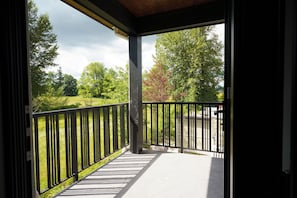 A covered outdoor area with a black metal railing, a concrete floor, and a view of a grassy field and trees.