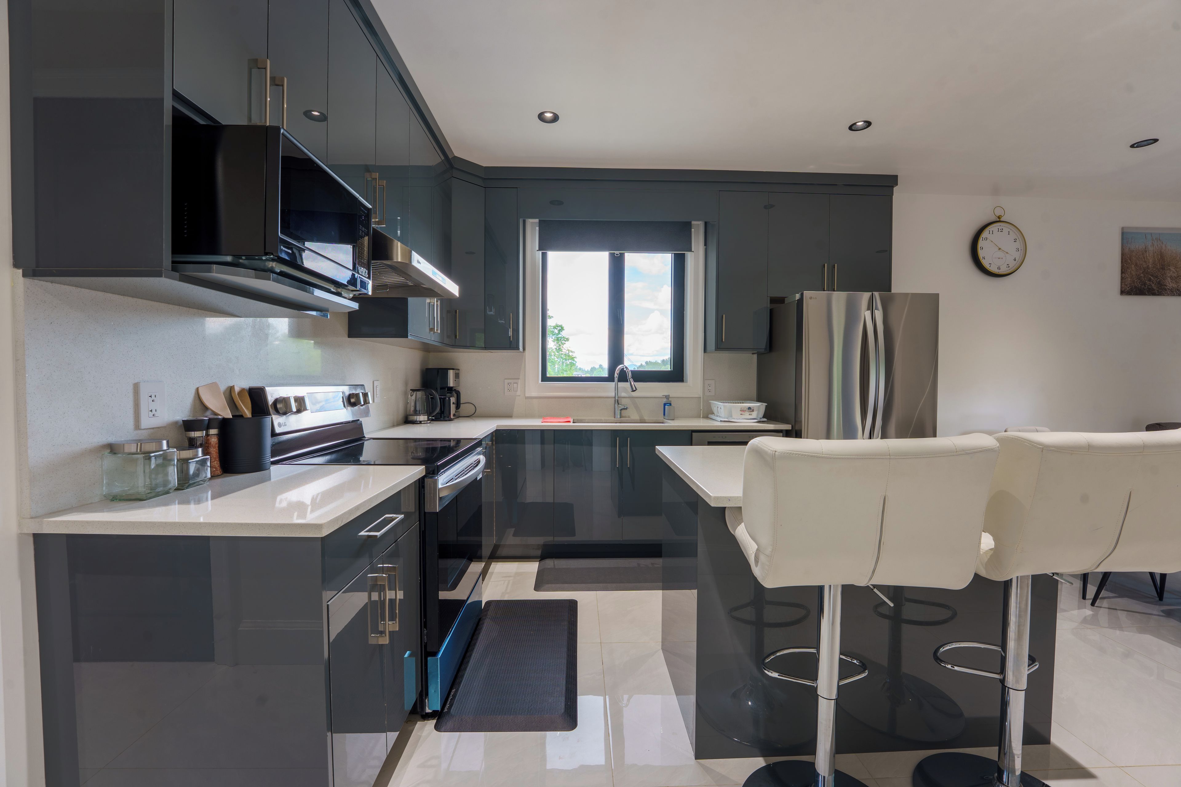 A modern kitchen with dark cabinetry, stainless appliances, a large window, and a bar area with stools.