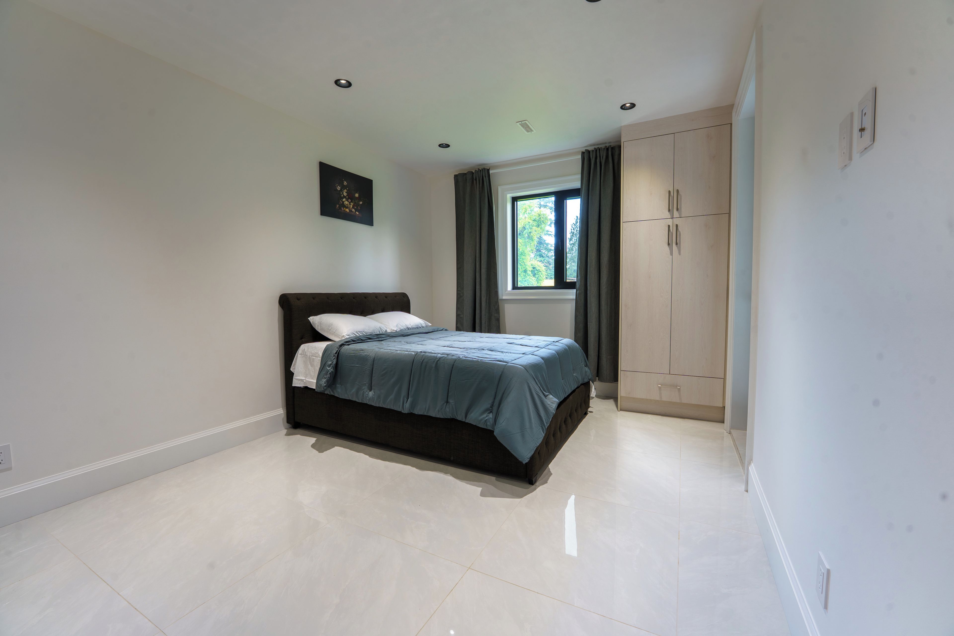 A bedroom with a bed, a window with curtains, a wardrobe, and recessed ceiling lights.