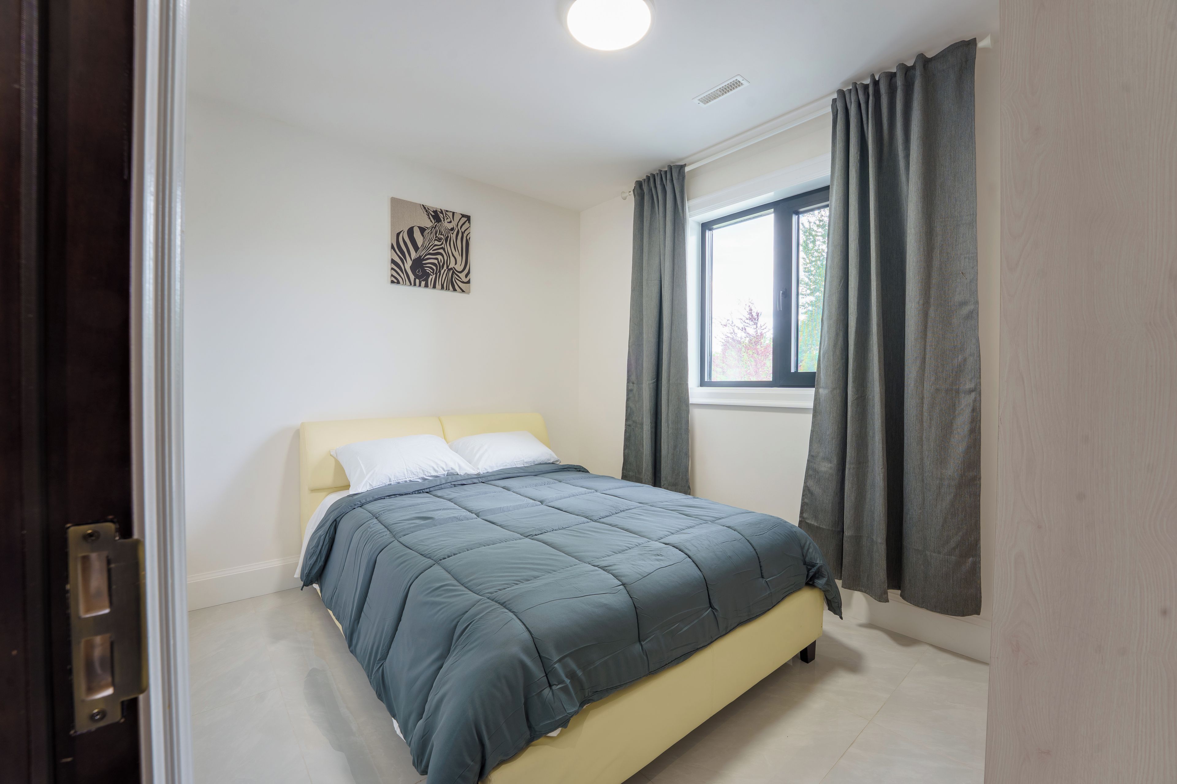 A bedroom with a bed, a window with curtains, a door, and a wall-mounted artwork.