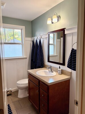 Un baño con un lavabo blanco, tocador y espejo. Hay toallas colgadas en un perchero, una ventana con vistas a árboles, y una puerta que da a otra habitación.