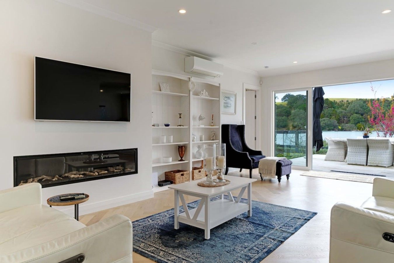 A modern living room with a fireplace, a flat-screen TV, a dining area with a table and chairs, and a view of a lake through large windows.