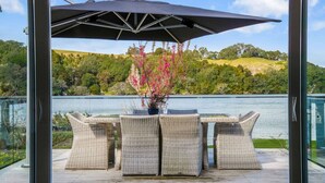 Terraza exterior con mobiliario de mimbre y una mesa, con vista a un lago y colinas al fondo.