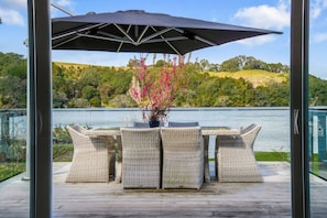Terraza exterior con mobiliario de mimbre y una mesa, con vista a un lago y colinas al fondo.