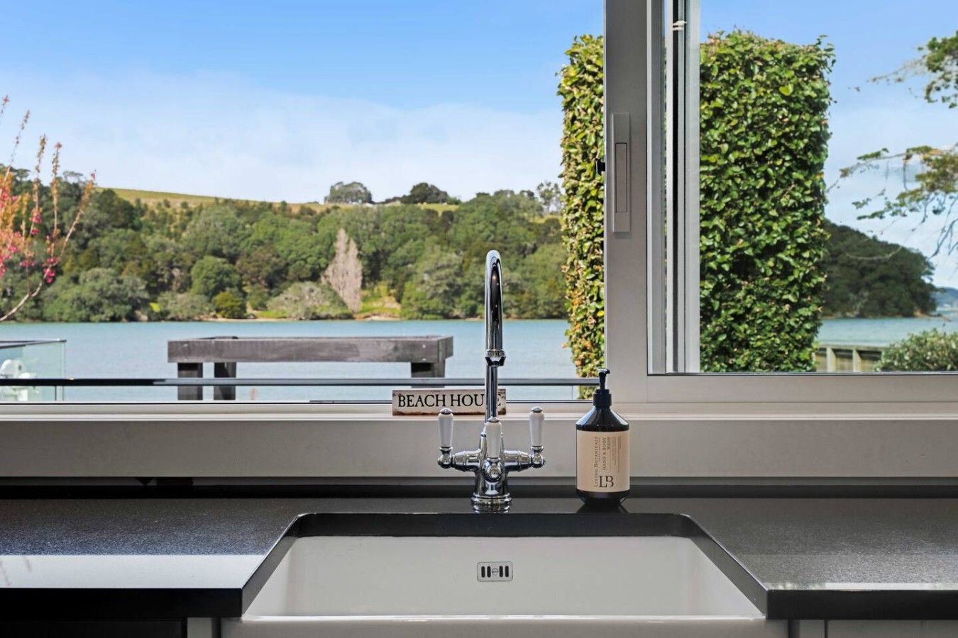 Eine Küchenarbeitsfläche mit Spüle, Wasserhahn und einer Handseifenflasche. Im Hintergrund Blick auf einen See und ein Schild mit der Aufschrift 'BEACH HOUSE'.