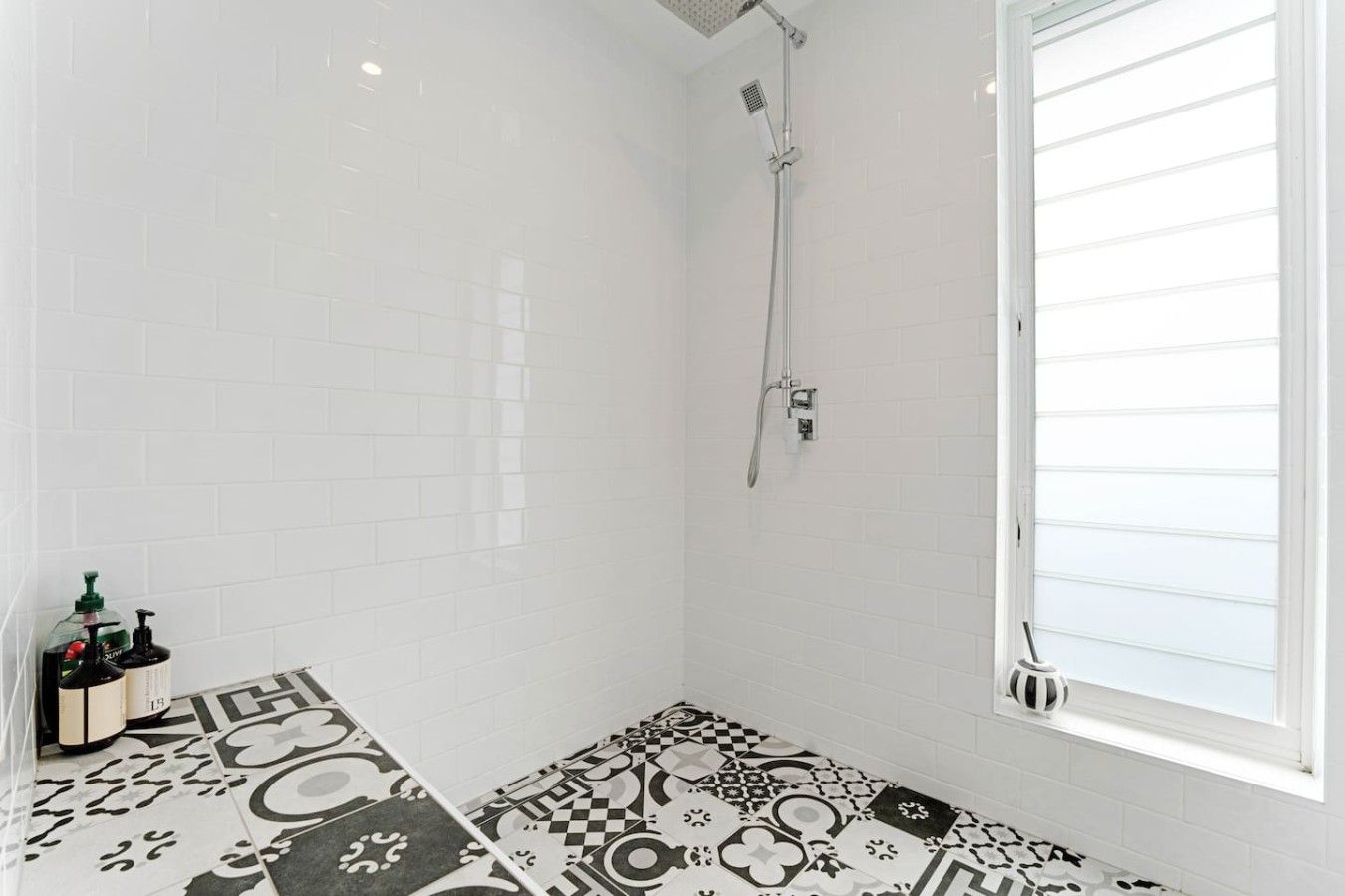 Een badkamer met witte betegelde wanden, een douche met een handdouche en vloertegels met een patroon.